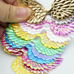May include: A collection of iridescent wing-shaped embellishments in various colors, including gold, silver, pink, blue, green, and yellow. The wings have a layered design, with each layer showcasing a different hue. These decorative elements are suitable for crafts and DIY projects.