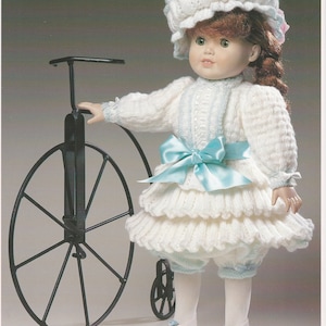 May include: A doll in a white knitted dress with a light blue satin bow and hat, next to a black metal bicycle. The image has the word "Eloisa" at the top. The doll's outfit is a knitted design.