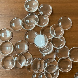 Könnte beinhalten: Eine Ansammlung klarer, runder Glascabochons auf einer Holzoberfläche. Die Cabochons haben eine gewölbte Oberfläche und sind in verschiedenen Größen erhältlich. Diese Glasteile eignen sich ideal für Schmuckherstellung und Bastelarbeiten.