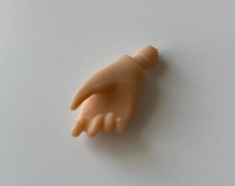 Może przedstawiać: Mała, beżowa plastikowa dłoń z lekko zgiętymi palcami. Ta miniaturowa dłoń jest prawdopodobnie elementem lalki lub figurki. Palce są szczegółowe, a ogólny wygląd jest gładki.