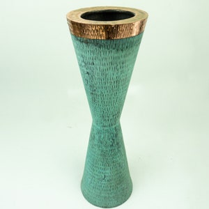 May include: A tall, hourglass-shaped teal vase with a textured surface. The top rim is accented with a copper band. The vase is a decorative item, suitable for displaying flowers or as a standalone art piece.