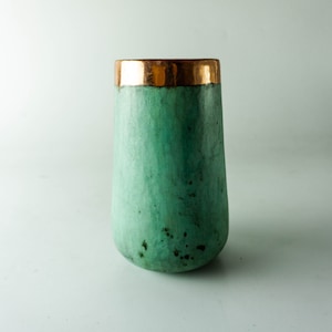 Könnte beinhalten: Eine hohe, zylindrische Vase mit abgerundeter Basis. Die Vase ist in einem verwitterten, meergrünen Farbton mit einem metallisch goldenen Rand bemalt. Die Vase hat eine glatte, matte Oberfläche und steht vor einem weißen Hintergrund.