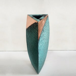 May include: A striking vase with a distinctive design. The top and a section are copper-toned, contrasting with the teal, textured body. The vase has a geometric form and a contemporary style, suitable for home decor.