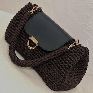 Puede incluir: Un bolso de mano marrón tejido a ganchillo con solapa negra y herrajes dorados. El bolso tiene un asa superior y una bandolera. El bolso mide aproximadamente 25 cm de ancho y 20 cm de alto.