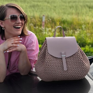 May include: A woven backpack with a flap closure and a top handle. The backpack is a light brown color with a matching flap and strap. The background features a field and a person wearing sunglasses and a pink shirt.