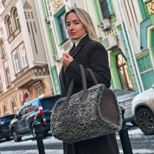 Może przedstawiać: Kobieta trzyma dużą, teksturowaną, szaro-brązową torebkę z dwoma uchwytami. Torba ma cylindryczny kształt i wydaje się być wykonana z miękkiego, tkanego materiału. Kobieta ma na sobie ciemnobrązowy płaszcz na ulicy miasta.