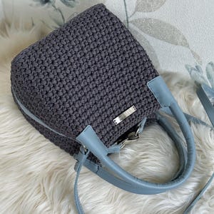 May include: A dark gray crocheted handbag with a structured square shape, featuring light blue faux leather handles and an adjustable shoulder strap. A small rectangular silver-toned metal plate is attached to the front of the textured bag.