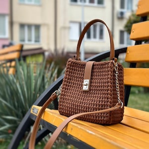 May include: A brown crochet handbag with a leather handle and strap. The bag has a rectangular shape with a flap closure and a gold-coloured metal clasp. The bag is resting on a wooden bench.