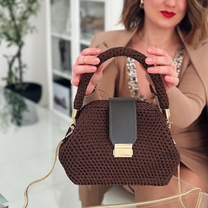 以下が含まれることがあります： 茶色の編み物ハンドバッグ。編み込みのハンドルとゴールドのチェーンストラップが付いています。バッグは、ゴールドの留め金が付いた濃いグレーのフラップが特徴です。バッグは人が持っています。