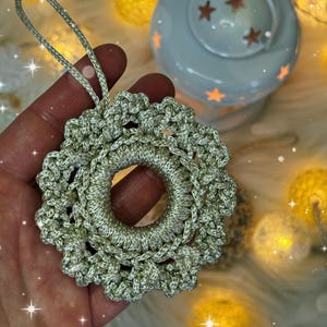 May include: A silver crocheted Christmas ornament with a circular design and a loop for hanging. The ornament is held in a hand, with a white lantern and string lights in the background. The ornament has a metallic sheen.