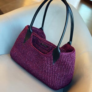 May include: A woven, plum-colored tote bag with black leather handles. The bag has a rectangular shape and a structured design. The handles are curved and attached to the bag with black leather accents. The bag is sitting on a light-colored surface.