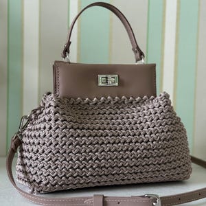 Könnte beinhalten: Eine handgefertigte, taupefarbene Handtasche mit gewebter Textur und strukturiertem Oberteil. Die Tasche hat einen Tragegriff und einen abnehmbaren Schulterriemen. Der obere Klappverschluss ist silberfarben. Die Tasche steht auf einer weißen Oberfläche.