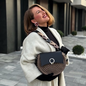 Könnte beinhalten: Eine schwarze und braune Umhängetasche mit rundem gewebtem Boden und schwarzem Lederklappe. Die Tasche hat einen silberfarbenen Ringverschluss und einen Kettenriemen mit braunen und schwarzen Gliedern. Die Person trägt einen weißen Mantel und schwarze Handschuhe.