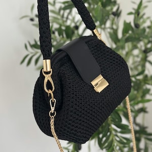 May include: A black crochet handbag with a braided handle and a gold chain strap. The bag features a black leather flap with a gold clasp. The bag is held up by a hand.