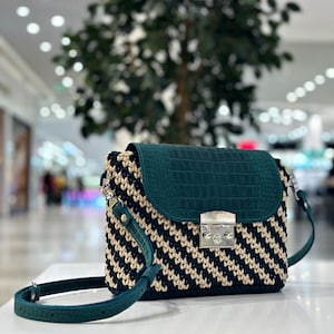 May include: A handcrafted handbag with a dark green crocodile-textured flap and strap. The body of the bag features a black and beige woven pattern. The bag has a silver-colored clasp.
