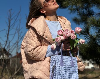 Puede incluir: Un bolso de mano de ganchillo azul claro con una correa para el hombro a juego. El bolso se sostiene frente a una persona que lleva una chaqueta acolchada rosa claro y sostiene un ramo de tulipanes rosas. El fondo es un cielo azul brillante y árboles.