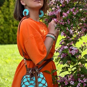 Pode incluir: Vestido laranja com cinto castanho e bolsa de croch&ecirc; a condizer com interior turquesa e sobreposi&ccedil;&atilde;o castanha. Os brincos s&atilde;o turquesa e castanhos, e as pulseiras s&atilde;o em tons neutros. O fundo apresenta uma &aacute;rvore florida e relva verde.