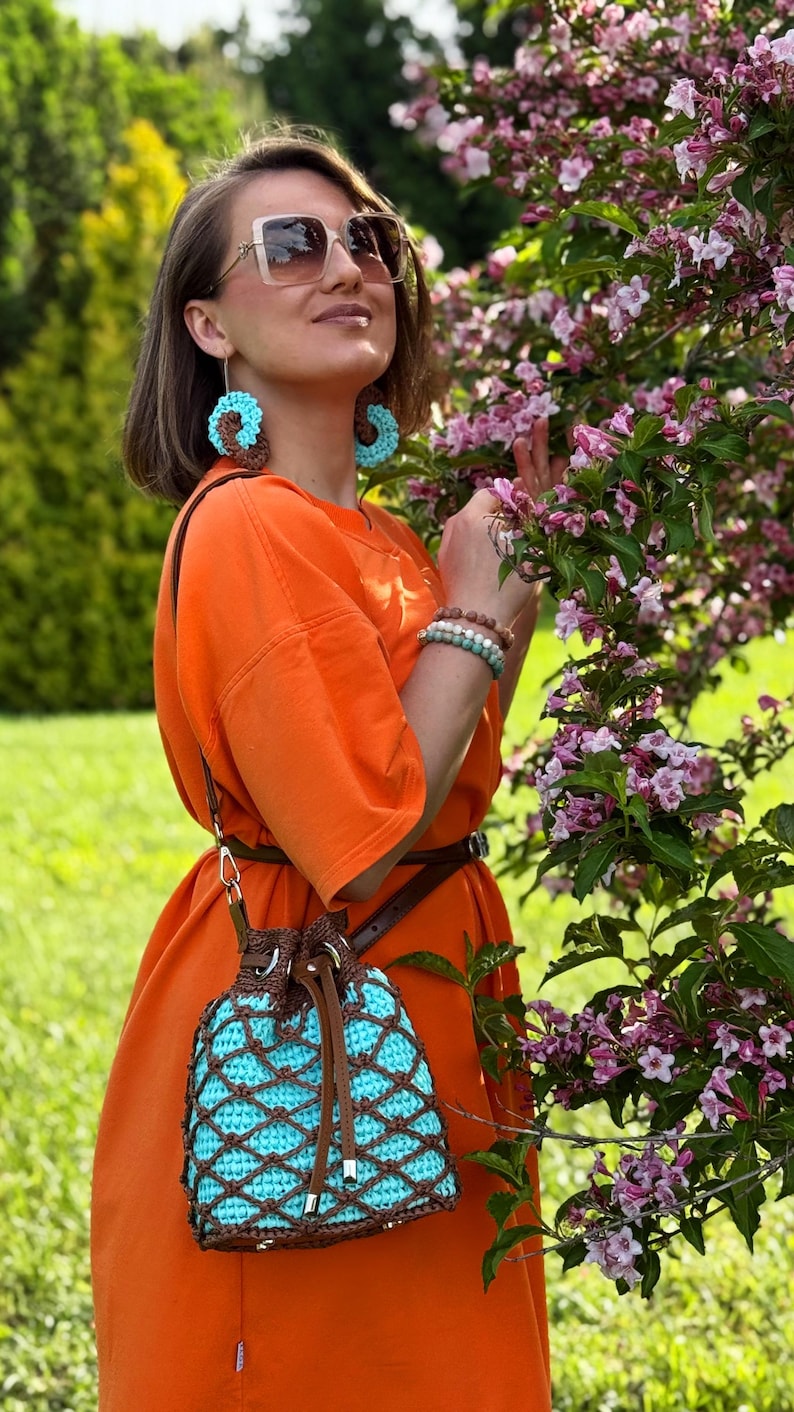 Pode incluir: Vestido laranja com cinto castanho e bolsa de croch&ecirc; a condizer com interior turquesa e sobreposi&ccedil;&atilde;o castanha. Os brincos s&atilde;o turquesa e castanhos, e as pulseiras s&atilde;o em tons neutros. O fundo apresenta uma &aacute;rvore florida e relva verde.