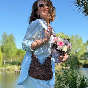 May include: A dark brown crochet shoulder bag with a scalloped top edge and a matching strap, worn over a light blue linen shirt. The bag features a dense, textured floral-like pattern created by intricate yarn loops and knots.