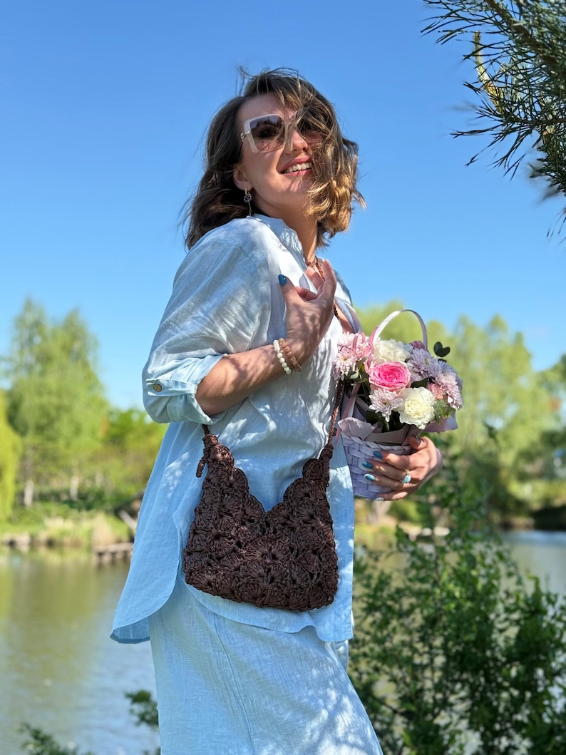 May include: A dark brown crochet shoulder bag with a scalloped top edge and a matching strap, worn over a light blue linen shirt. The bag features a dense, textured floral-like pattern created by intricate yarn loops and knots.