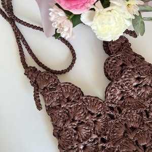 May include: A dark brown crocheted bag featuring a repeating shell stitch pattern and a long, braided shoulder strap. The bag is constructed from a glossy, ribbon-like material and displays a textured, openwork design throughout the body.