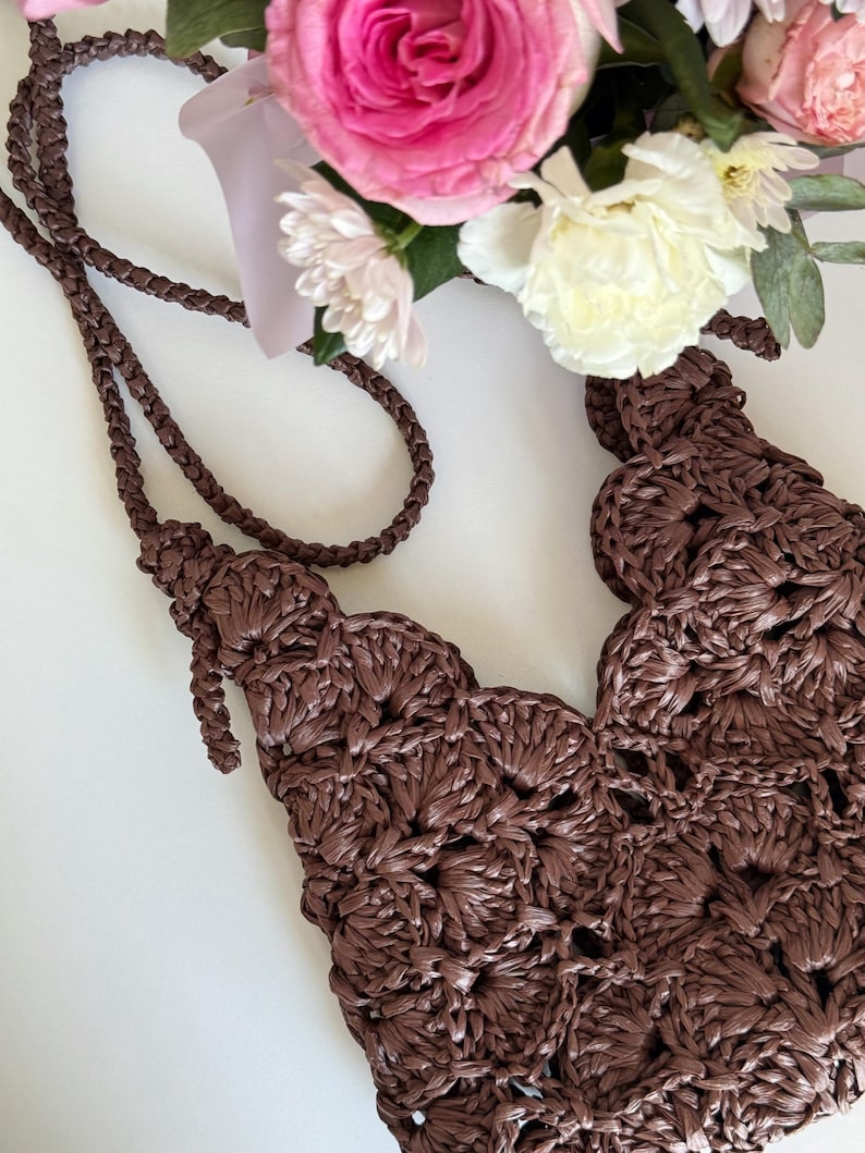 May include: A dark brown crocheted bag featuring a repeating shell stitch pattern and a long, braided shoulder strap. The bag is constructed from a glossy, ribbon-like material and displays a textured, openwork design throughout the body.