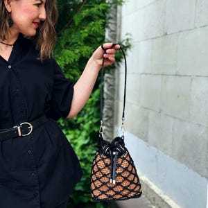 Pode incluir: Uma bolsa de balde de croch&ecirc; preta e castanha com fecho de cord&atilde;o e uma longa al&ccedil;a preta. A bolsa apresenta um padr&atilde;o de diamante e ferragens prateadas. A bolsa &eacute; segurada por uma pessoa.