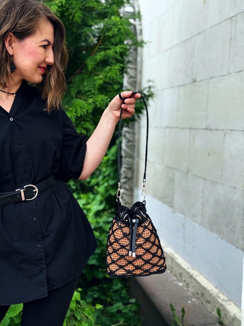 Pode incluir: Uma bolsa de balde de croch&ecirc; preta e castanha com fecho de cord&atilde;o e uma longa al&ccedil;a preta. A bolsa apresenta um padr&atilde;o de diamante e ferragens prateadas. A bolsa &eacute; segurada por uma pessoa.