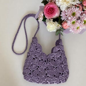 May include: A lavender crochet handbag featuring a repeating shell stitch pattern and a matching braided shoulder strap. The bag has a scalloped top edge and is constructed from a shiny, textured yarn, showcasing a detailed openwork design.