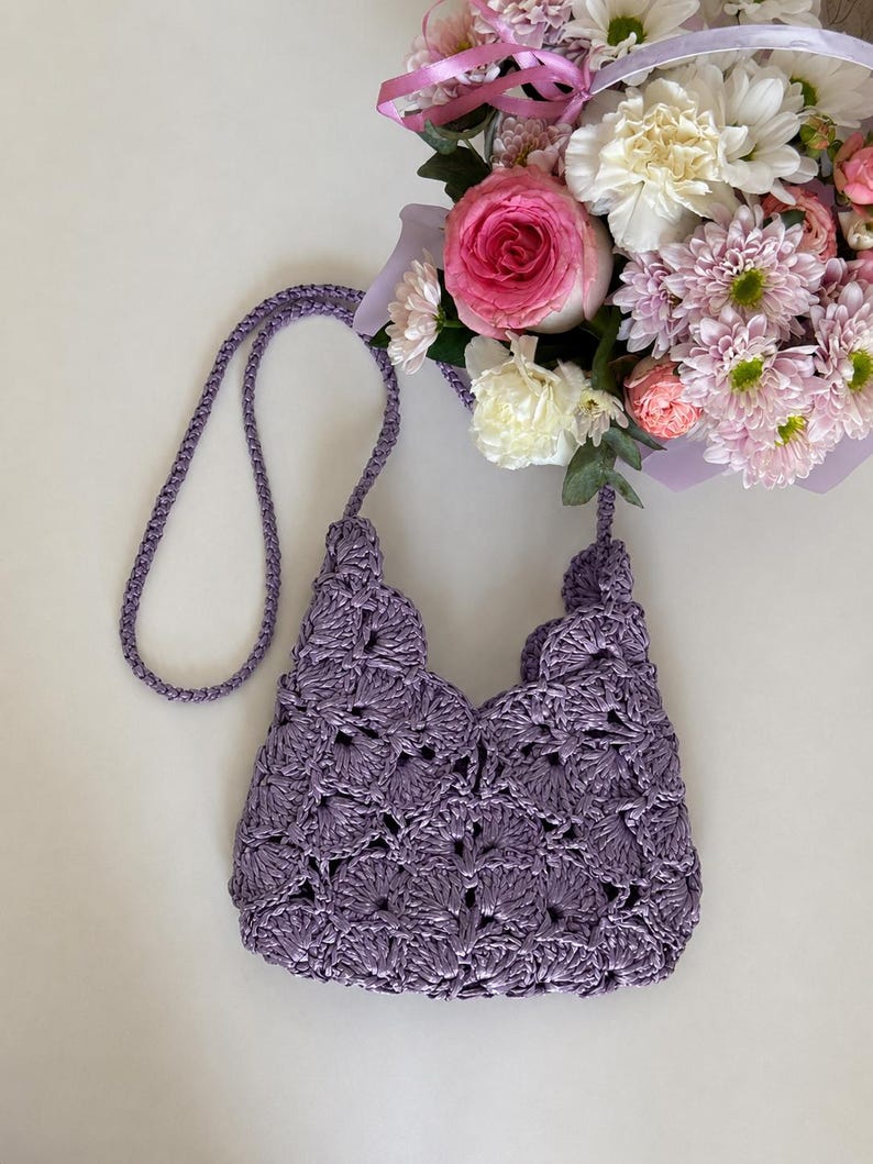 May include: A lavender crochet handbag featuring a repeating shell stitch pattern and a matching braided shoulder strap. The bag has a scalloped top edge and is constructed from a shiny, textured yarn, showcasing a detailed openwork design.