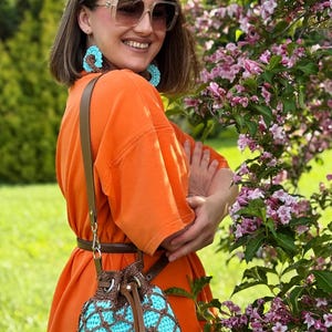 Pode incluir: Uma bolsa de balde de croch&ecirc; turquesa e marrom com fecho de cord&atilde;o e al&ccedil;a de couro marrom. A bolsa &eacute; usada no ombro. A pessoa est&aacute; usando um vestido laranja e brincos combinando.