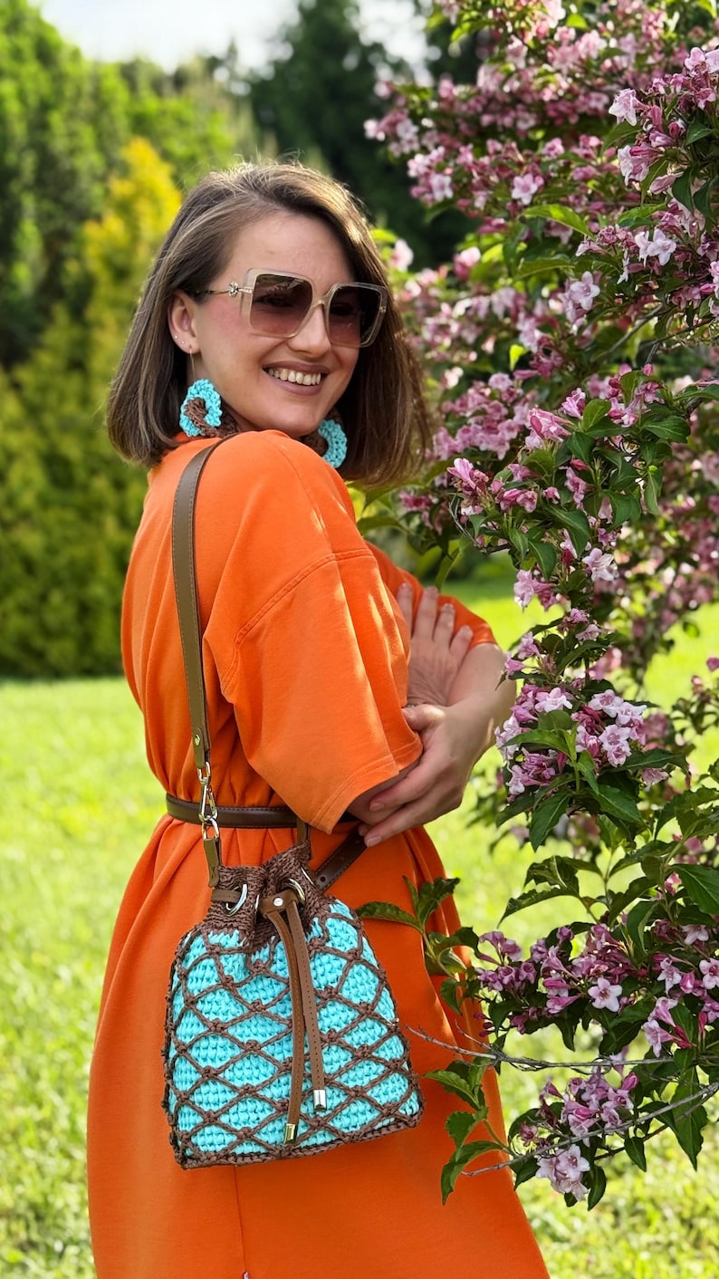 Pode incluir: Uma bolsa de balde de croch&ecirc; turquesa e marrom com fecho de cord&atilde;o e al&ccedil;a de couro marrom. A bolsa &eacute; usada no ombro. A pessoa est&aacute; usando um vestido laranja e brincos combinando.