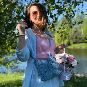 May include: A light blue crochet crossbody bag with a scalloped pattern, worn over a light blue button-down shirt and a pink tank top. The bag features a matching braided strap and is paired with a small woven basket containing a floral arrangement.
