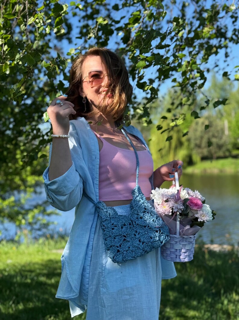 May include: A light blue crochet crossbody bag with a scalloped pattern, worn over a light blue button-down shirt and a pink tank top. The bag features a matching braided strap and is paired with a small woven basket containing a floral arrangement.