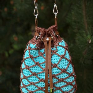 Pode incluir: Uma bolsa de balde de croch&ecirc; feita &agrave; m&atilde;o com interior turquesa e exterior tecido marrom. A bolsa apresenta al&ccedil;as de couro marrom e um fecho de cord&atilde;o. &Eacute; suspensa por duas al&ccedil;as de couro marrom com fechos de metal prateado.