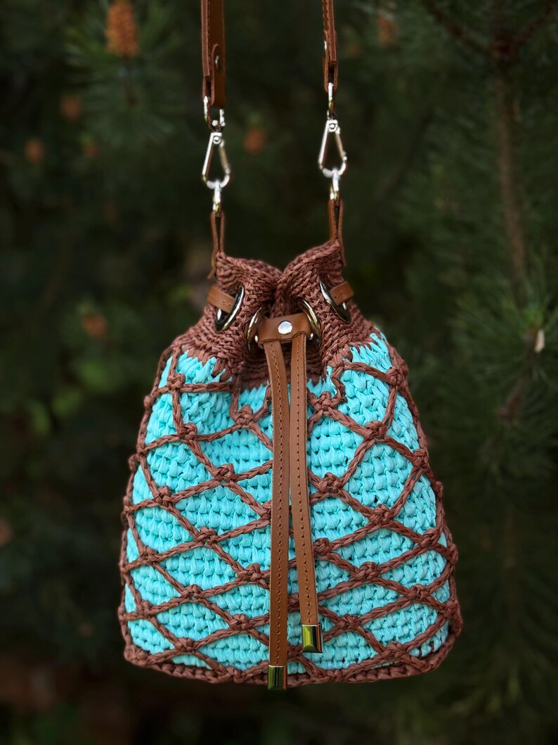 Pode incluir: Uma bolsa de balde de croch&ecirc; feita &agrave; m&atilde;o com interior turquesa e exterior tecido marrom. A bolsa apresenta al&ccedil;as de couro marrom e um fecho de cord&atilde;o. &Eacute; suspensa por duas al&ccedil;as de couro marrom com fechos de metal prateado.