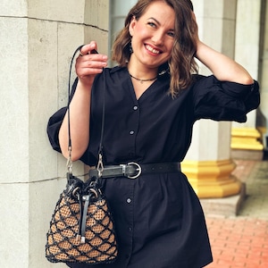 Pode incluir: Uma bolsa preta com formato de balde com um design tecido em padr&atilde;o de diamante em bege e preto. A bolsa tem um fecho de cord&atilde;o e uma al&ccedil;a preta. A bolsa &eacute; segurada por uma pessoa que usa uma camisa e um cinto pretos.