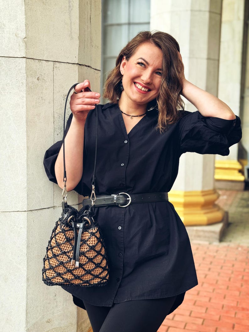 Pode incluir: Uma bolsa preta com formato de balde com um design tecido em padr&atilde;o de diamante em bege e preto. A bolsa tem um fecho de cord&atilde;o e uma al&ccedil;a preta. A bolsa &eacute; segurada por uma pessoa que usa uma camisa e um cinto pretos.
