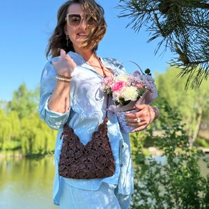 May include: A dark brown crocheted crossbody bag with a textured floral pattern and a thin strap. The bag features a scalloped top edge and a zipper closure, crafted from a dark brown yarn with a visible woven structure and intricate detailing.