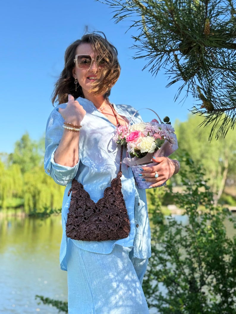 May include: A dark brown crocheted crossbody bag with a textured floral pattern and a thin strap. The bag features a scalloped top edge and a zipper closure, crafted from a dark brown yarn with a visible woven structure and intricate detailing.