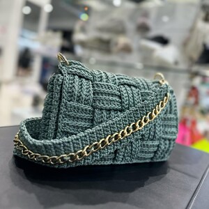 May include: A dark sage green handbag featuring a woven crochet texture. The bag has a structured rectangular shape and includes a gold-toned metal chain strap attached to the sides with gold hardware, providing a metallic contrast to the yarn.