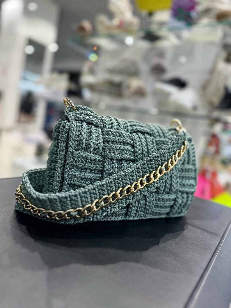 May include: A dark sage green handbag featuring a woven crochet texture. The bag has a structured rectangular shape and includes a gold-toned metal chain strap attached to the sides with gold hardware, providing a metallic contrast to the yarn.