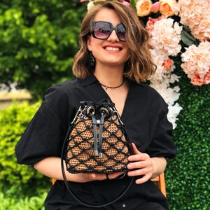 Pode incluir: Bolsa de balde tecida preta e castanha com fecho de cord&atilde;o e al&ccedil;a de ombro preta. A bolsa tem um padr&atilde;o geom&eacute;trico e ferragens prateadas. A bolsa &eacute; segurada por uma pessoa que usa uma camisa preta e &oacute;culos de sol.