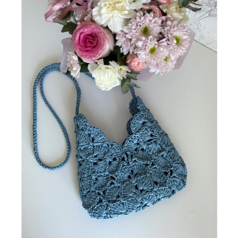 May include: A dusty blue crochet handbag featuring a scalloped shell pattern throughout the body. The bag includes a long, thin matching crochet shoulder strap and an open top design, showcasing a textured, woven construction in a solid blue hue.