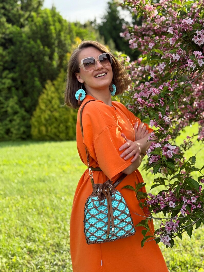 Pode incluir: Um vestido laranja com cinto castanho e uma bolsa de croch&ecirc; turquesa e castanha com fecho de cord&atilde;o. A bolsa tem uma al&ccedil;a de couro castanho. A pessoa est&aacute; usando brincos turquesa e &oacute;culos de sol.