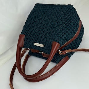 May include: A dark teal crochet handbag with brown leather handles and straps. The bag has a rectangular shape with a rounded top and a small metal tag. The leather handles are curved and attached to the bag.