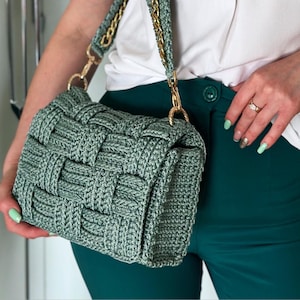 May include: A sage green handbag featuring a woven crochet texture. The bag includes a matching crochet strap reinforced with a gold-toned metal chain, connected to the main body by gold-toned metal lobster clasps on each side of the rectangular frame.