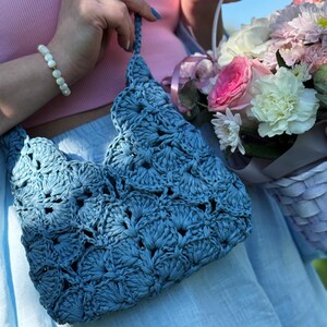 May include: A dusty blue crochet handbag featuring a repeating scalloped shell pattern. The bag has a matching braided shoulder strap and is constructed from a textured, matte material. The design consists of interconnected fan-shaped stitches.