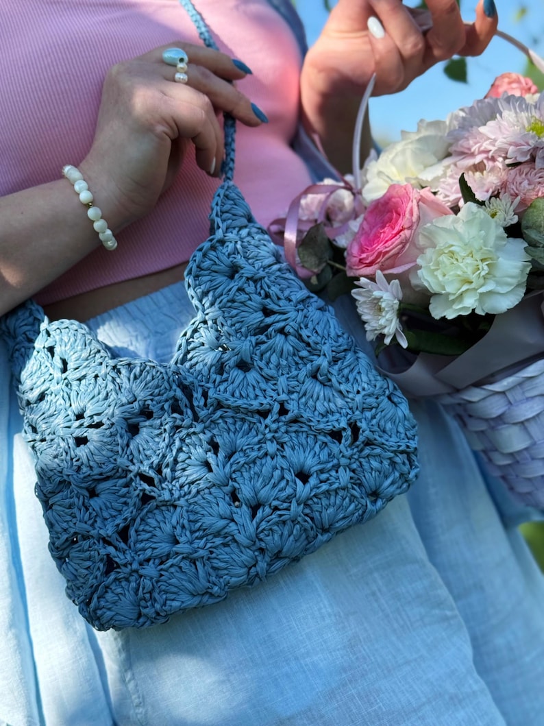 May include: A dusty blue crochet handbag featuring a repeating scalloped shell pattern. The bag has a matching braided shoulder strap and is constructed from a textured, matte material. The design consists of interconnected fan-shaped stitches.