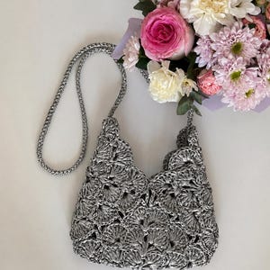 May include: A metallic silver crochet handbag featuring a repeating fan-like shell stitch pattern throughout the body. The bag includes a matching braided silver shoulder strap and an open top design, showcasing a textured, shiny finish.